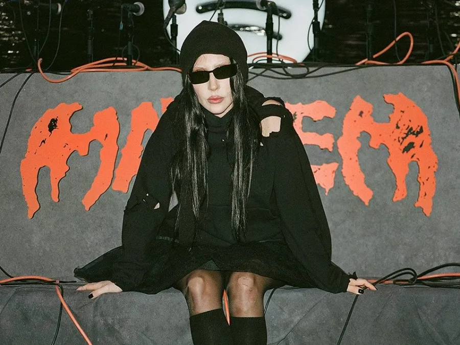 Η Lady Gaga με μαύρο σκούφο και γυαλιά ηλίου κάθεται μπροστά από ένα πορτοκαλί λογότυπο.