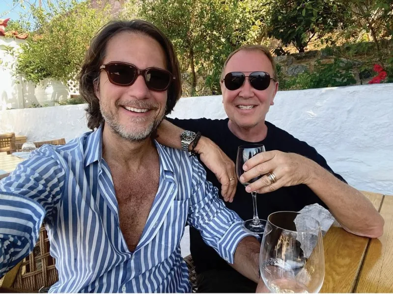 Ο Michael Kors και ένας άνδρας κάθονται σε ένα τραπέζι σε εξωτερικό χώρο.