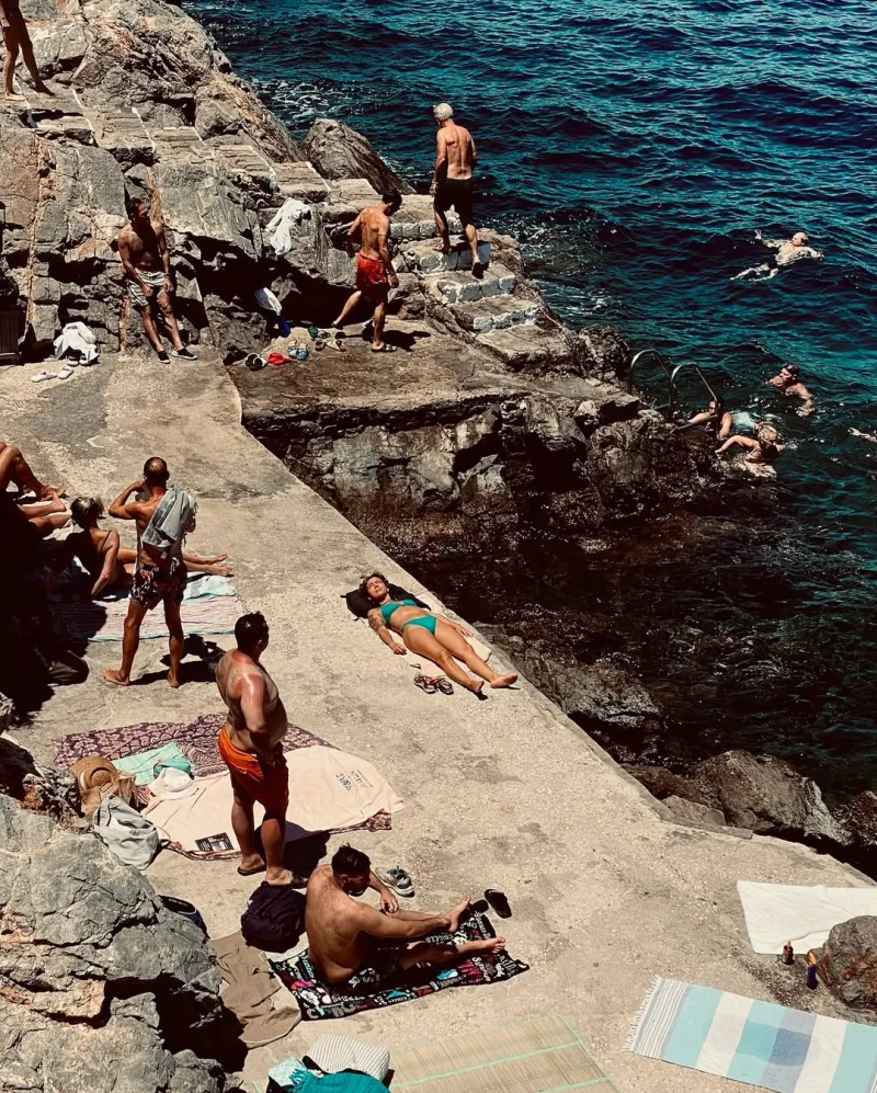 Άνθρωποι απολαμβάνουν το μπάνιο και την ηλιοθεραπεία σε βραχώδη ακτή στην Ελλάδα.