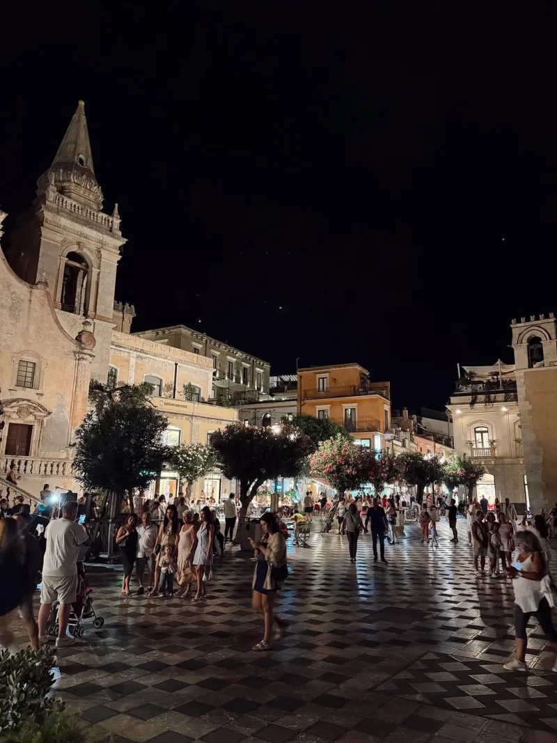 Νυχτερινή άποψη πλατείας με κόσμο και ιστορικά κτίρια.