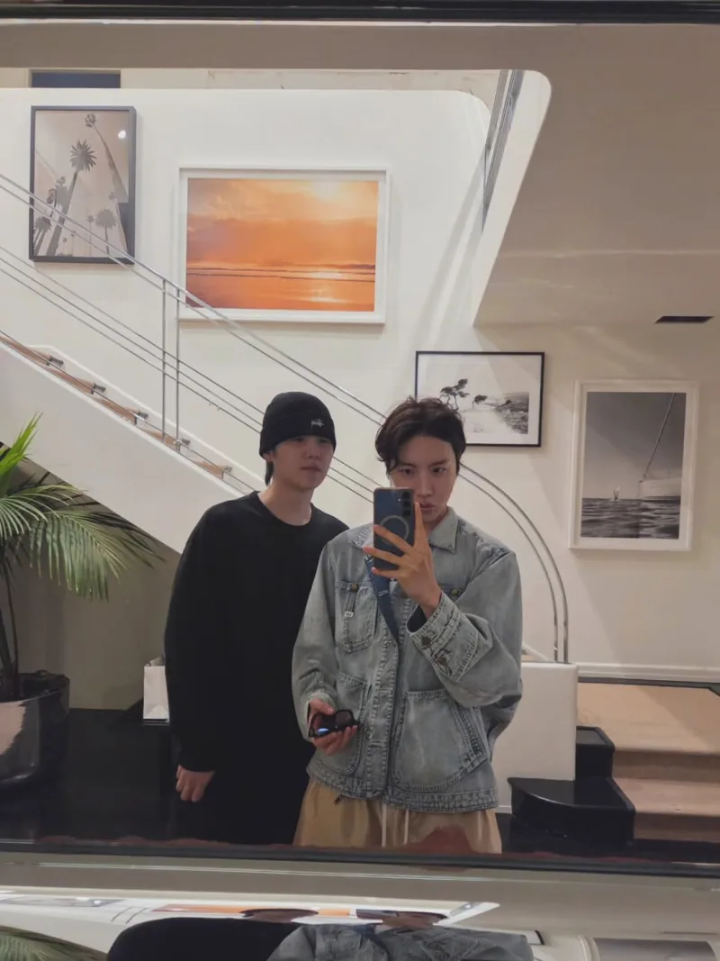Δύο μέλη των BTS σε selfie μπροστά από καθρέφτη σε εσωτερικό χώρο με κάδρα.