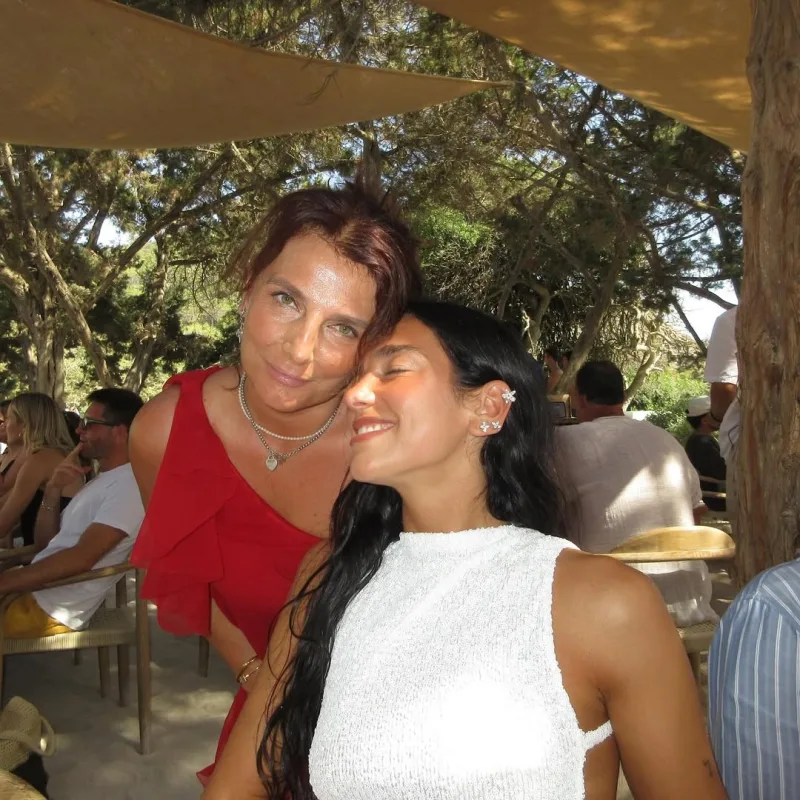 Η Dua Lipa με λευκό φόρεμα δίπλα σε γυναίκα με κόκκινο φόρεμα σε εξωτερικό χώρο.