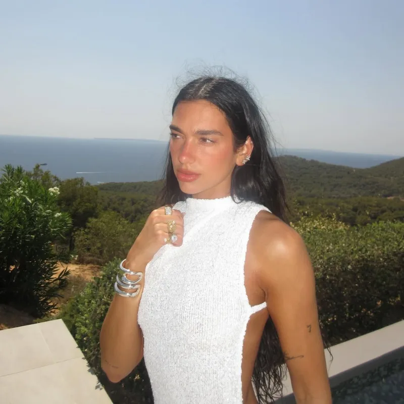 Η Dua Lipa με λευκό εξώπλατο φόρεμα σε εξωτερικό χώρο με θέα τη θάλασσα.