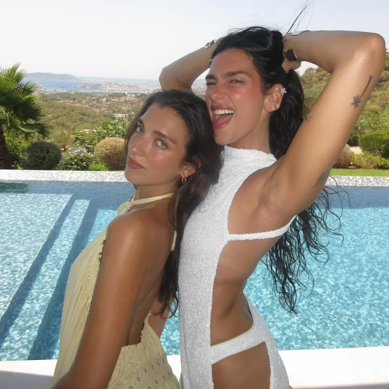 Η Dua Lipa με λευκό cut-out φόρεμα δίπλα σε φίλη της μπροστά από πισίνα.