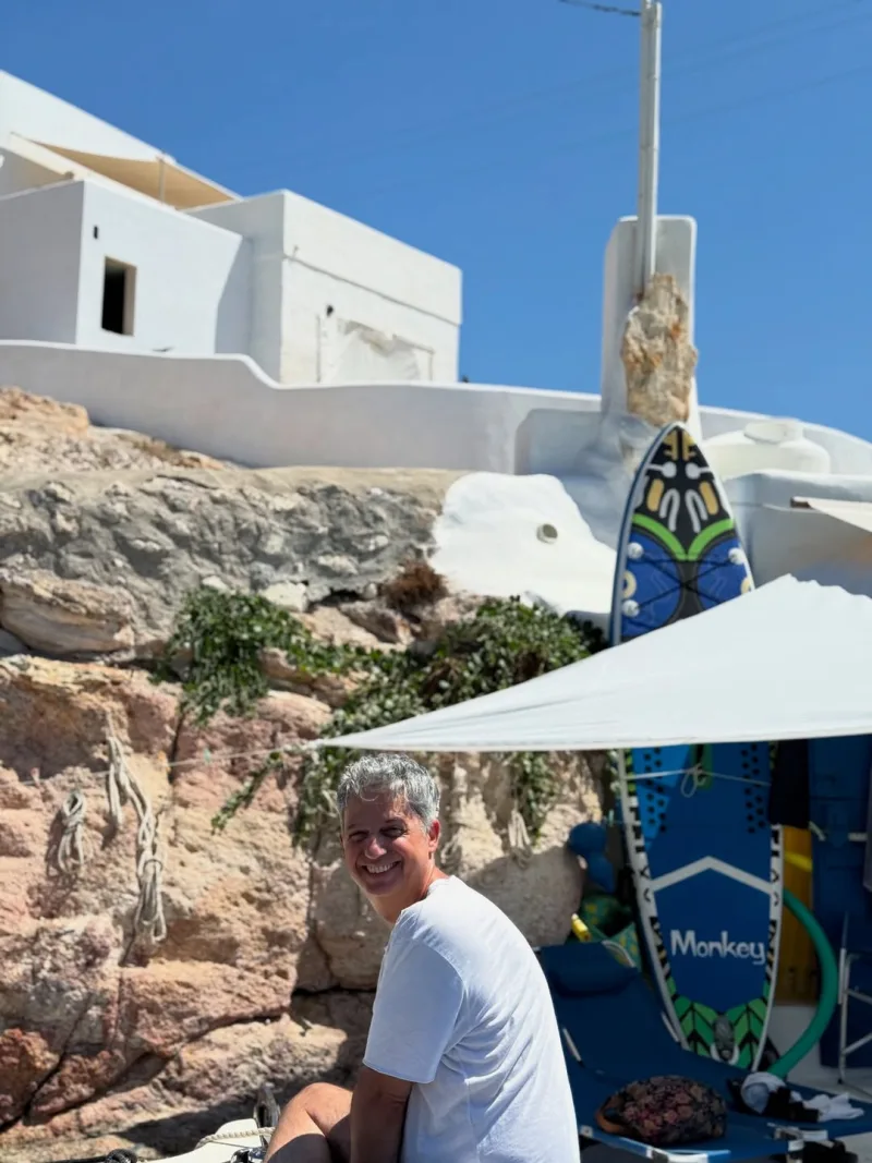 Ένας άνδρας με γκρίζα μαλλιά χαμογελάει μπροστά από λευκά κυκλαδίτικα σπίτια και μια σανίδα surf.