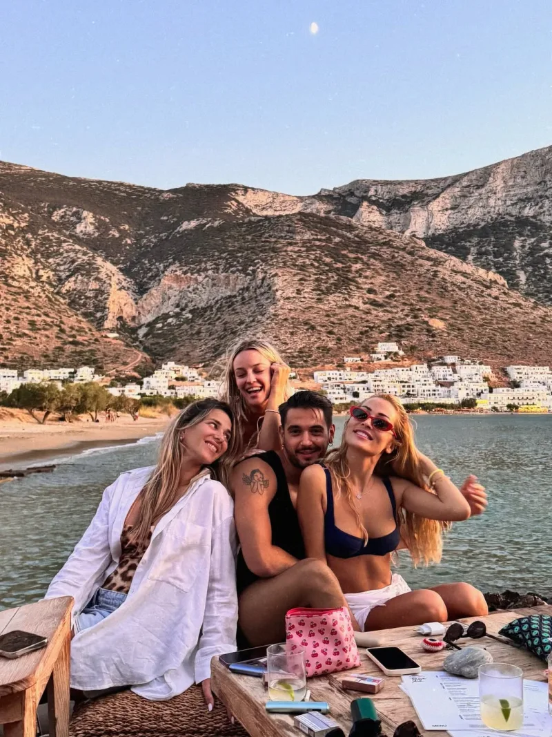 Παρέα φίλων σε παραθαλάσσιο τραπέζι με φόντο βουνό και οικισμό.