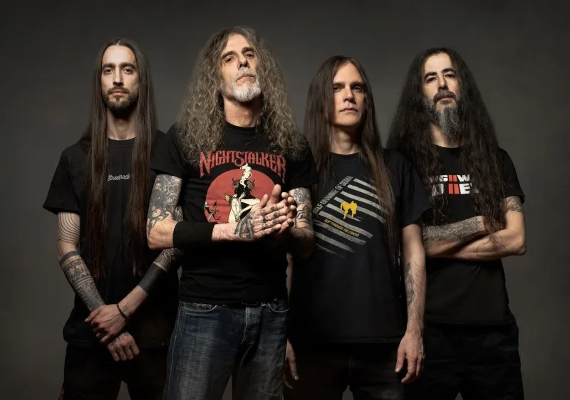 Τέσσερις άνδρες με μακριά μαλλιά και μαύρα t-shirts ποζάρουν σε γκρίζο φόντο.