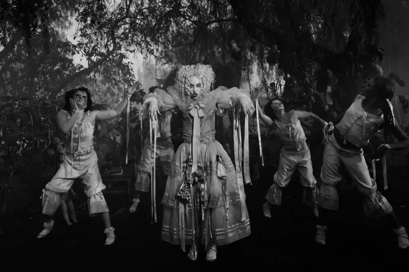 Η Lady Gaga με λευκό φόρεμα και μακιγιάζ κούκλας περιτριγυρισμένη από χορευτές σε ασπρόμαυρο σκηνικό.