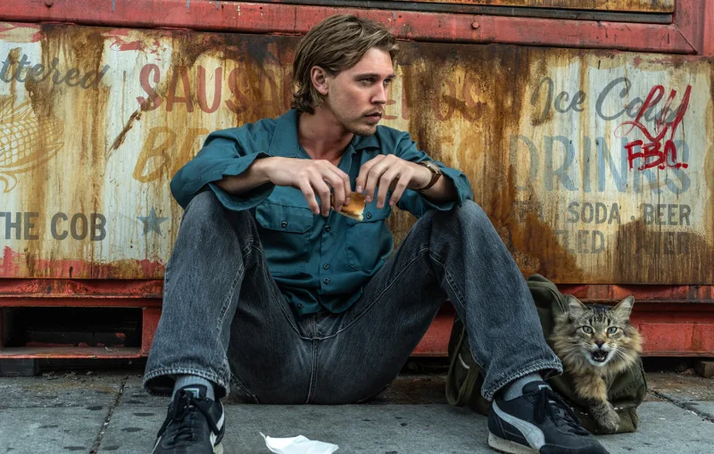 Ο Austin Butler κάθεται στο έδαφος μπροστά από έναν σκουριασμένο τοίχο με μια γάτα δίπλα του.