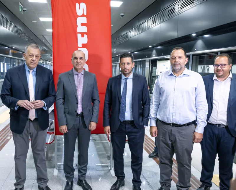 Πέντε άνδρες με κοστούμια και πουκάμισα ποζάρουν μπροστά από κόκκινο λάβαρο σε σταθμό μετρό.