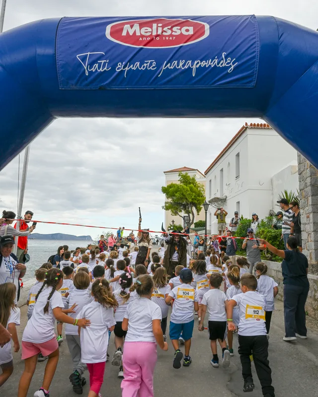 Παιδιά ξεκινούν αγώνα δρόμου κάτω από μπλε αψίδα στο Spetses Mini Marathon 2025.
