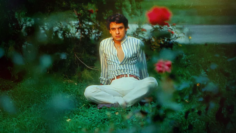 Ο Juan Gabriel σε νεαρή ηλικία κάθεται στο γρασίδι ανάμεσα σε τριαντάφυλλα.