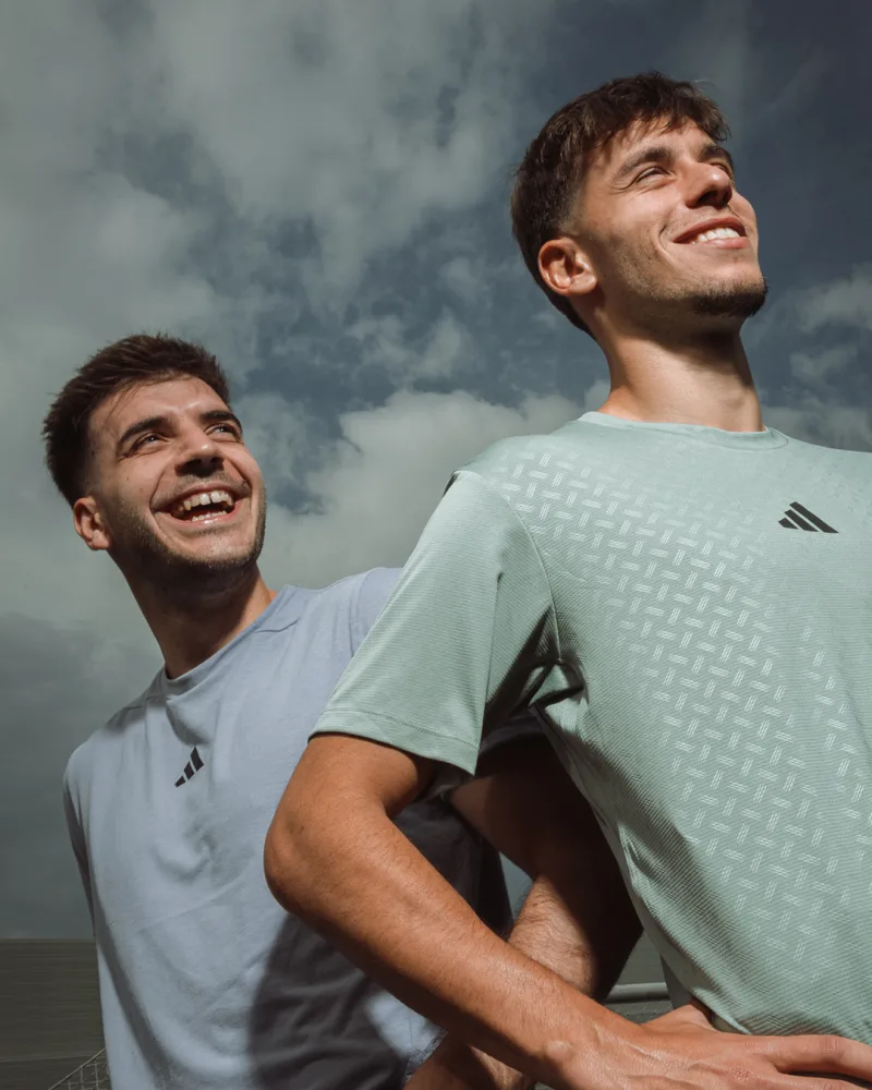 Δύο νεαροί άνδρες με αθλητικά μπλουζάκια adidas χαμογελούν κάτω από συννεφιασμένο ουρανό.