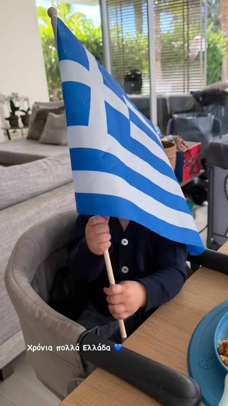 Παιδί κρατάει την ελληνική σημαία καθισμένο σε καρέκλα.