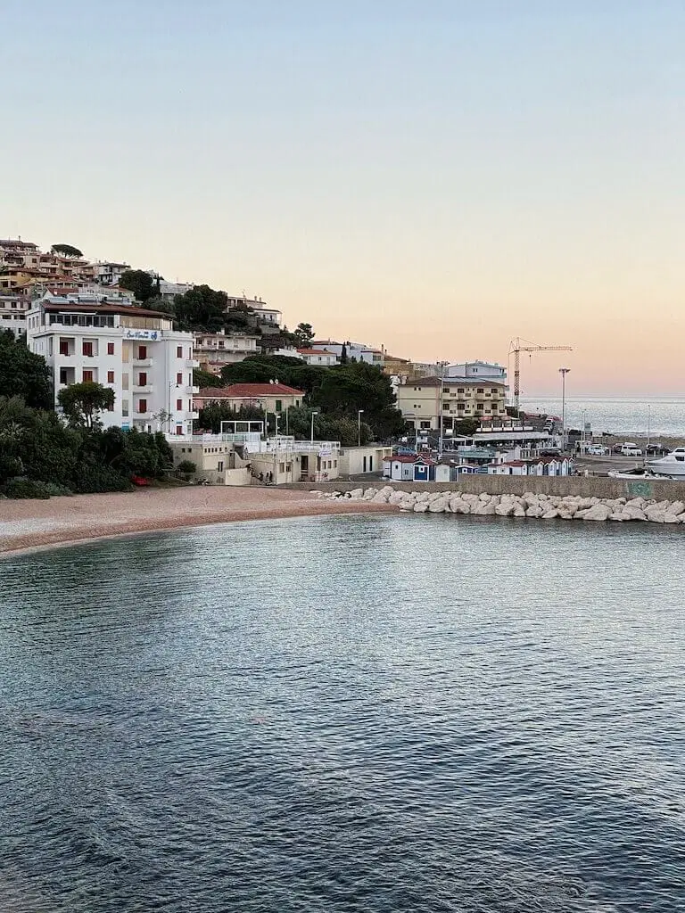Παραθαλάσσιος οικισμός στη Σαρδηνία με λευκά κτίρια και λιμάνι κατά το ηλιοβασίλεμα.