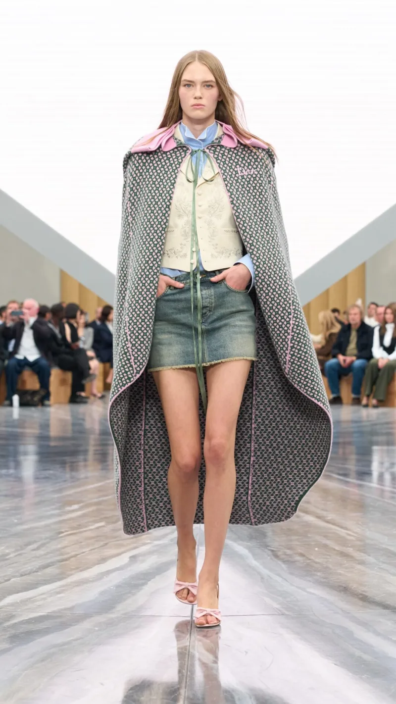 Μοντέλο στην επίδειξη Dior SS26 με μίνι τζιν φούστα και μακριά κάπα.