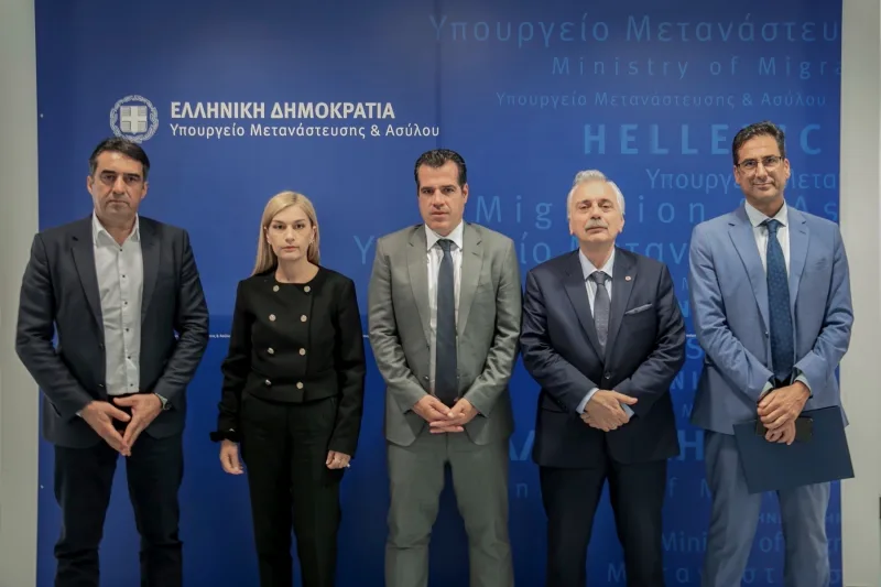 Πέντε άτομα σε επίσημη φωτογράφιση μπροστά από το λογότυπο του Υπουργείου Μετανάστευσης και Ασύλου.