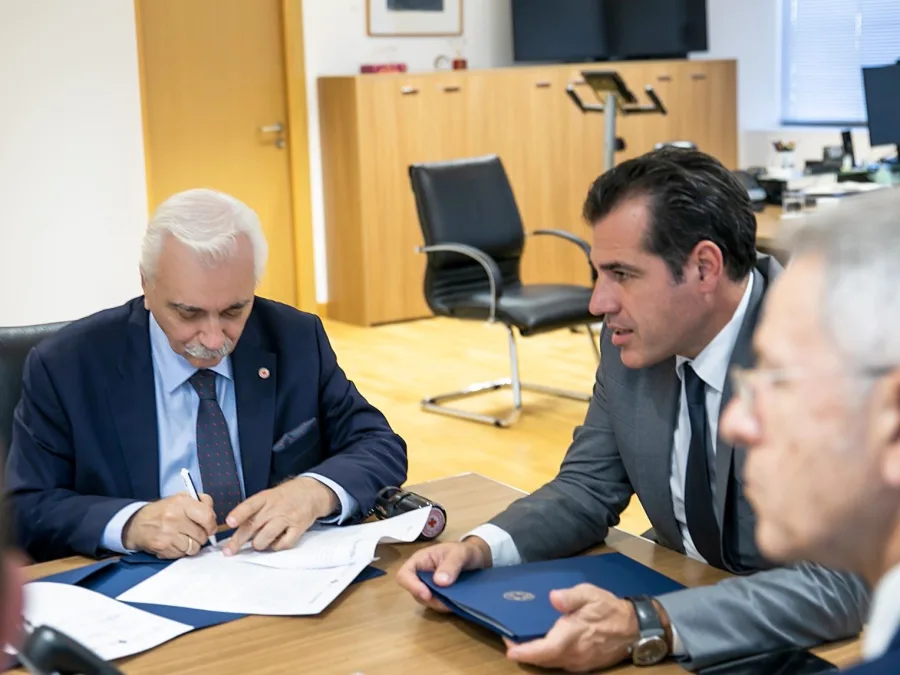 Δύο άνδρες με κοστούμια υπογράφουν έγγραφα σε γραφείο.