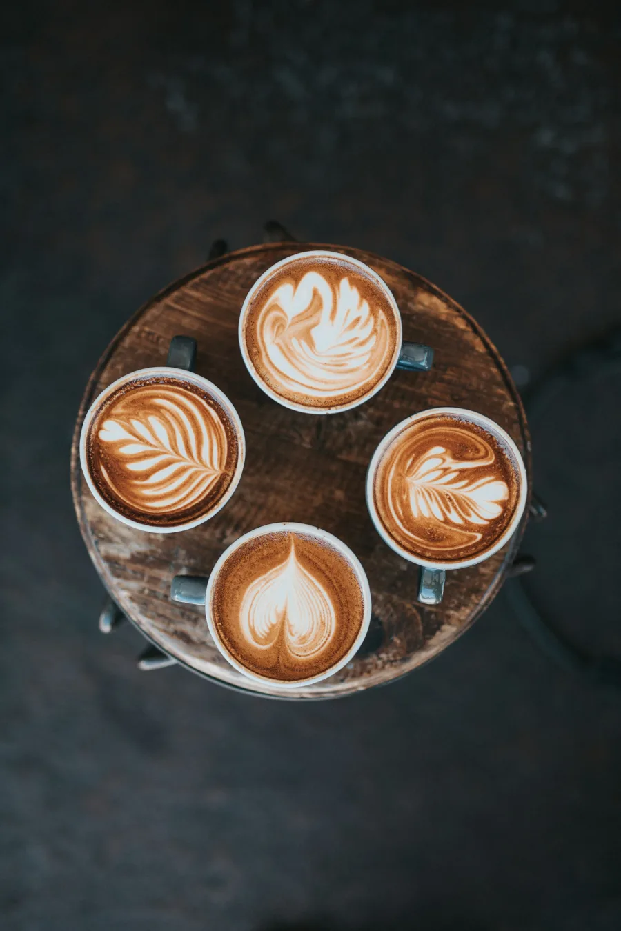 Τέσσερα φλιτζάνια καφέ latte με διαφορετικά σχέδια latte art πάνω σε ξύλινο σκαμπό.
