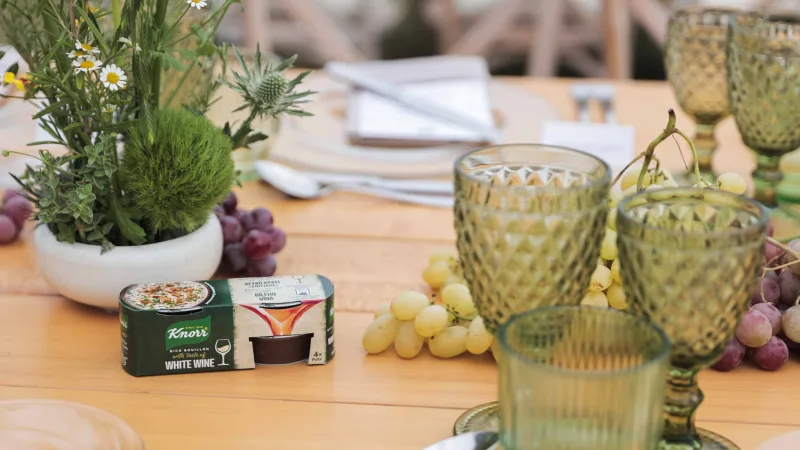 Συσκευασία Knorr Σπιτικός Ζωμός με λευκό κρασί σε στρωμένο τραπέζι με σταφύλια και ποτήρια.