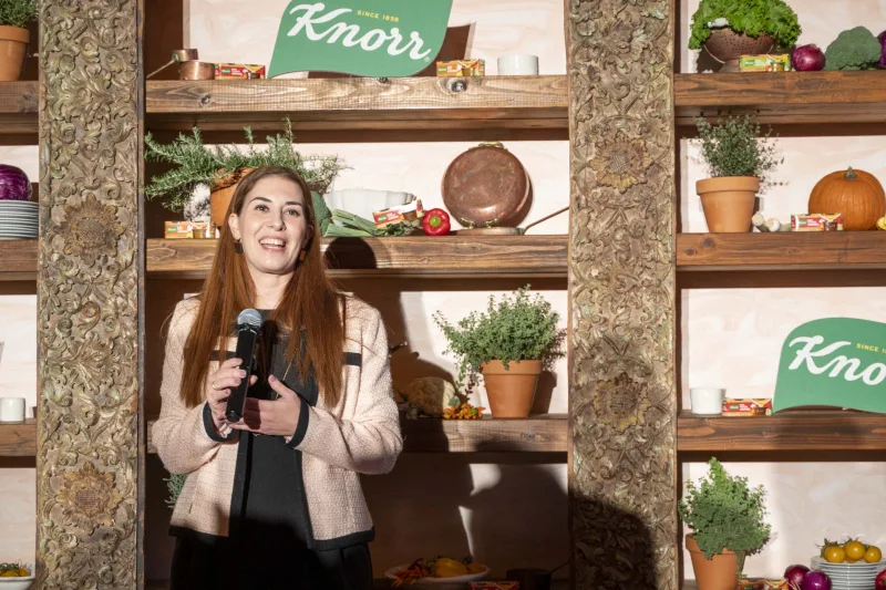 Γυναίκα μιλάει σε εκδήλωση της Knorr μπροστά από ράφια με υλικά μαγειρικής.
