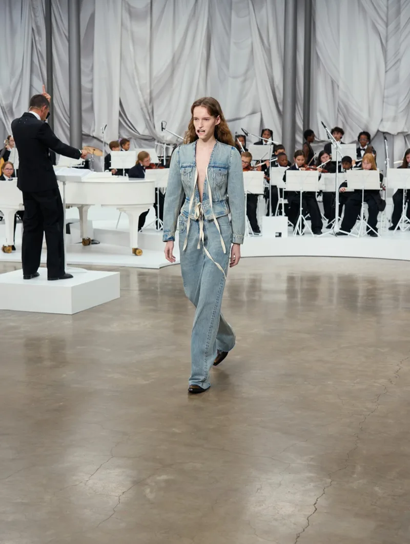 Μοντέλο με denim σύνολο Maison Margiela σε πασαρέλα με ορχήστρα.