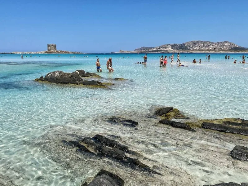 Παραλία στη Σαρδηνία με κρυστάλλινα νερά, βράχια και κόσμο να κολυμπά.