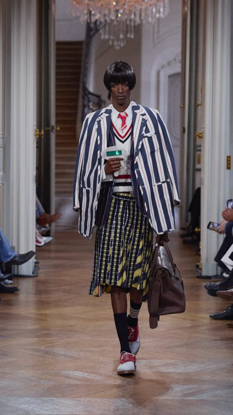 Μοντέλο στην επίδειξη Thom Browne SS26 με ριγέ σακάκι και πλισέ φούστα.