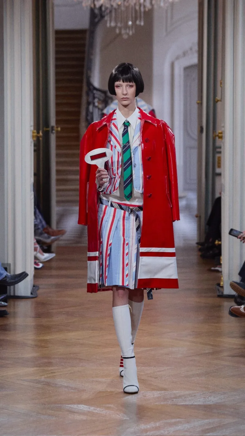 Μοντέλο στην επίδειξη Thom Browne SS26 με κόκκινο παλτό και ριγέ σύνολο.