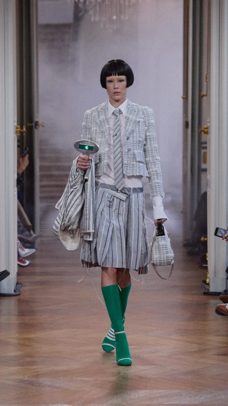 Μοντέλο στην επίδειξη Thom Browne SS26 με γκρι ριγέ φούστα και πράσινες μπότες.