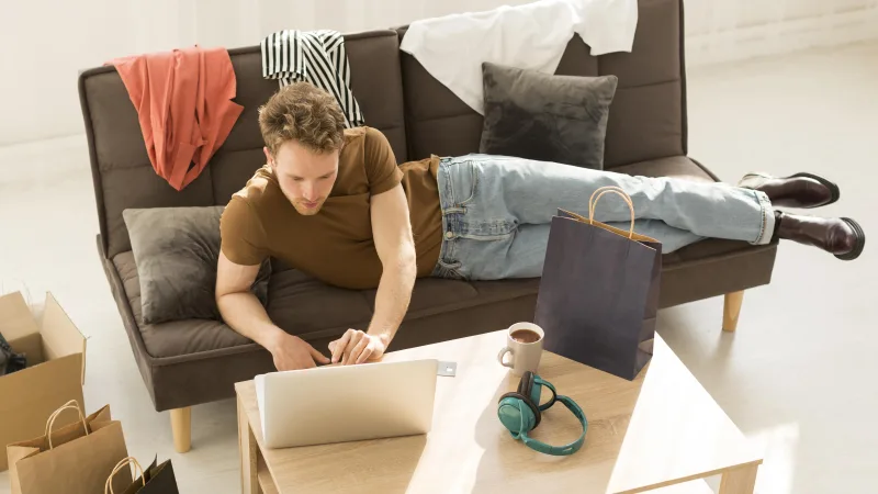 Νεαρός άνδρας χαλαρώνει στον καναπέ με laptop και ψώνια, αποφεύγοντας δραστηριότητες κατά τον Ανάδρομο Ερμή.