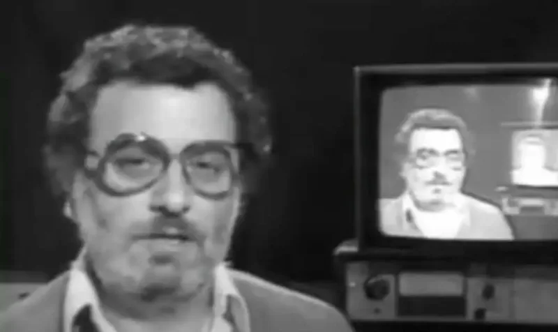 Παρουσιαστής με γυαλιά σε εκπομπή που άλλαξε την ελληνική τηλεόραση, με τον εαυτό του να εμφανίζεται στην οθόνη.
