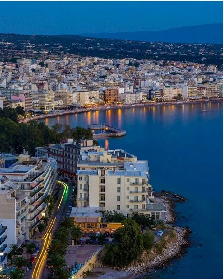 Πανοραμική άποψη της παραλιακής πόλης, ιδανική για μονοήμερη εκδρομή από την Αθήνα, με φωτισμένα κτίρια και θάλασσα.