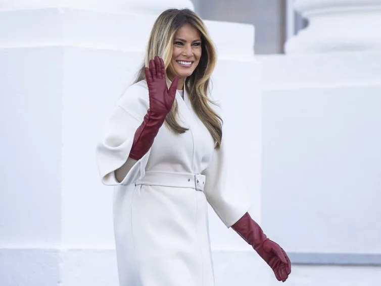 Η Melania Trump χαιρετάει φορώντας μακριά κόκκινα γάντια και λευκό παλτό έξω από τον Λευκό Οίκο.