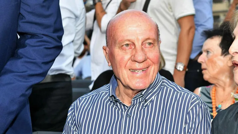 Ένας ηλικιωμένος άνδρας με ριγέ πουκάμισο χαμογελά σε εξωτερικό χώρο.