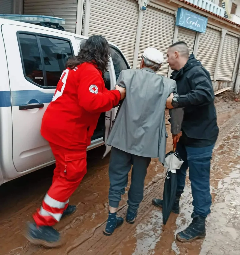 Εθελόντρια του Ελληνικού Ερυθρού Σταυρού βοηθά ηλικιωμένο άνδρα σε πλημμυρισμένο δρόμο.