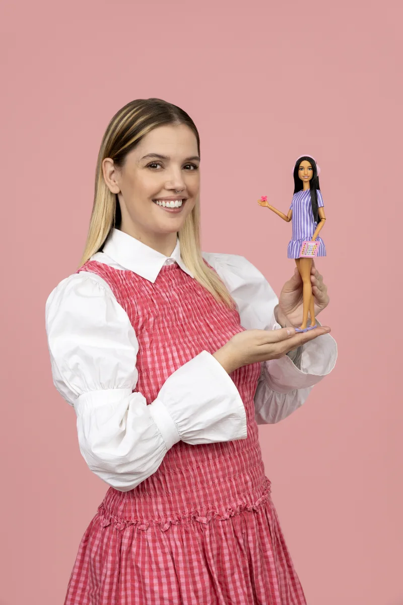 Μια γυναίκα κρατά την πρώτη κούκλα Barbie με αυτισμό σε ροζ φόντο.