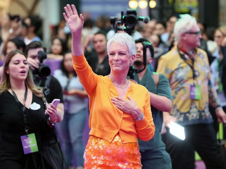 Η Jamie Lee Curtis χαιρετά το κοινό με ενθουσιασμό, φορώντας ένα πορτοκαλί ζακετάκι και φόρεμα με παγιέτες.