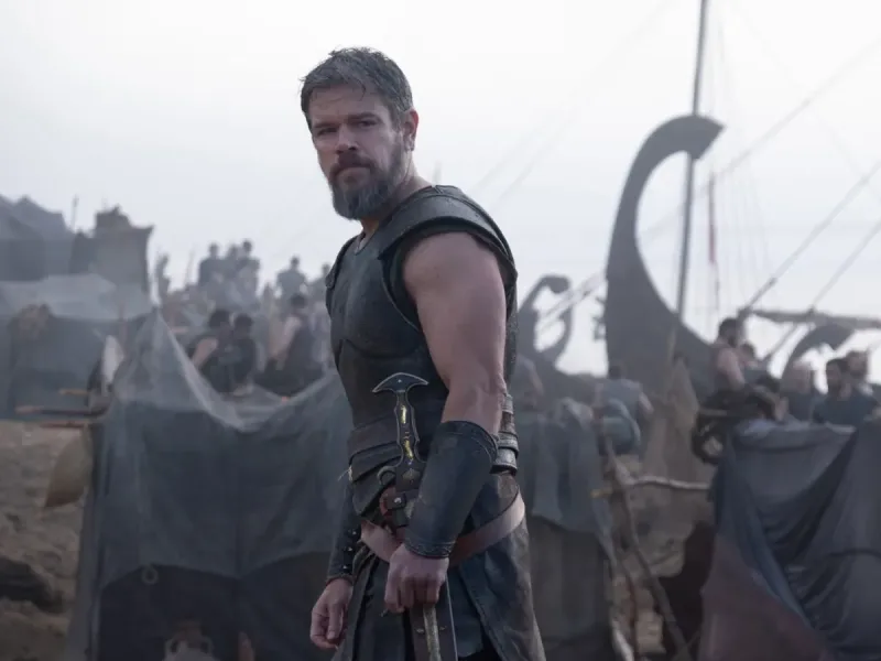 Ο Matt Damon ως στρατιώτης με πανοπλία και σπαθί σε σκηνικό της Οδύσσειας, με πλοία και στρατόπεδο στο φόντο.