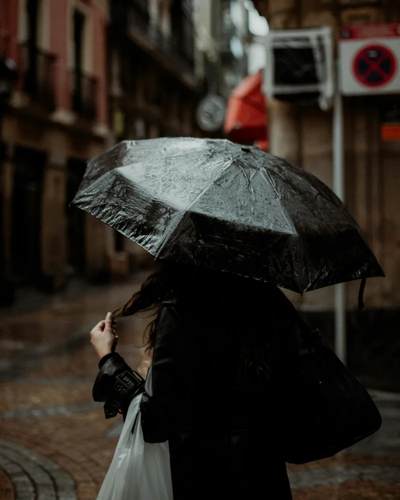 Γυναίκα με μαύρη ομπρέλα περπατά σε βρεγμένο δρόμο κατά τη διάρκεια βροχής.