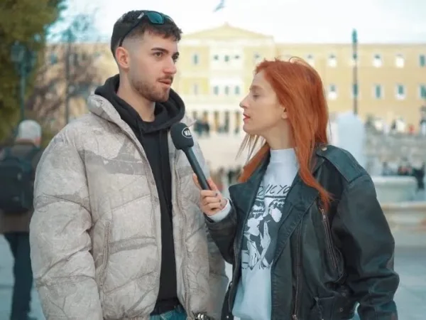 Μια γυναίκα με κόκκινα μαλλιά παίρνει συνέντευξη από έναν άνδρα με μπουφάν στο δρόμο.