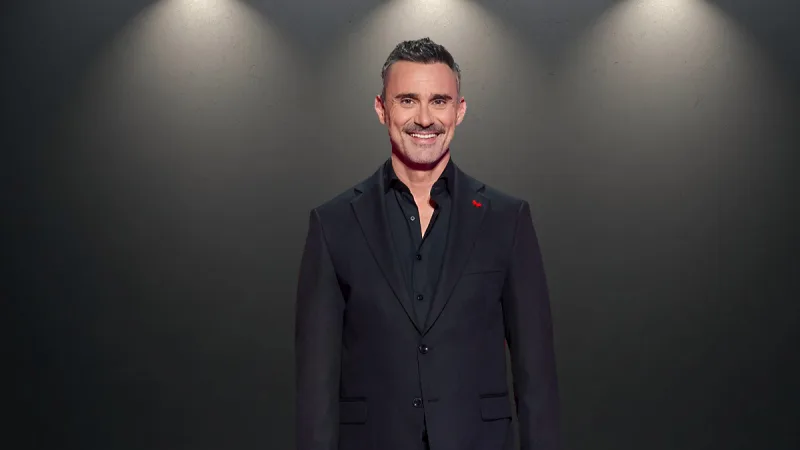 Ο παρουσιαστής του Α’ ημιτελικού της Eurovision 2026, Freddie, ποζάρει με κοστούμι μπροστά σε σκούρο φόντο.