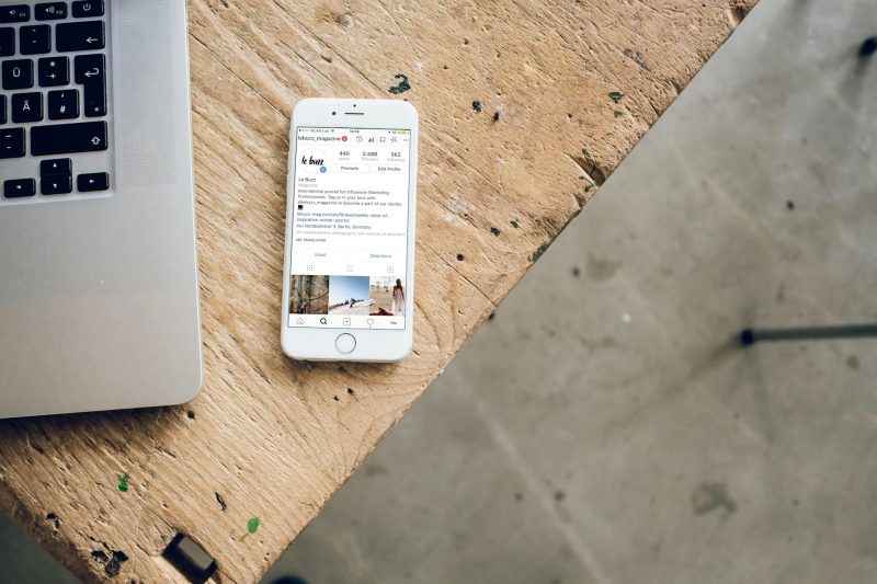 Ένα smartphone με ανοιχτό το Instagram δίπλα σε ένα laptop πάνω σε ξύλινο τραπέζι.