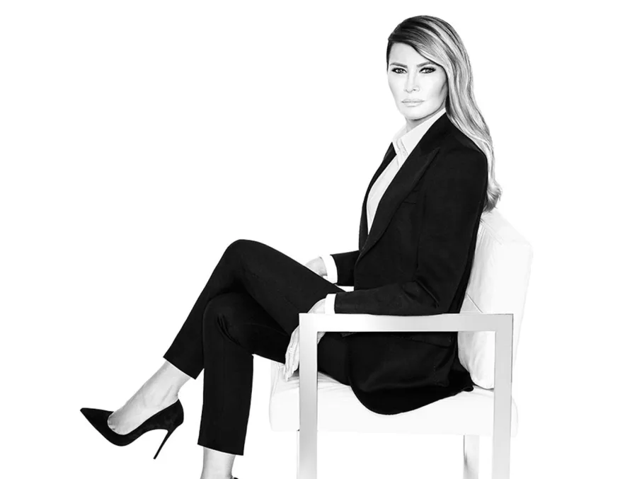 Η Melania Trump ποζάρει καθιστή σε καρέκλα φορώντας μαύρο κοστούμι.