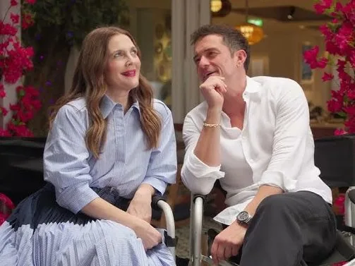 Η Drew Barrymore και ο Orlando Bloom κάθονται δίπλα-δίπλα και χαμογελούν.