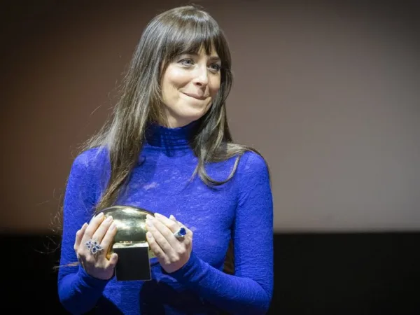 Η Dakota Johnson κρατάει ένα χρυσό βραβείο, φορώντας ένα μπλε φόρεμα με μακριά μανίκια και εντυπωσιακά δαχτυλίδια.