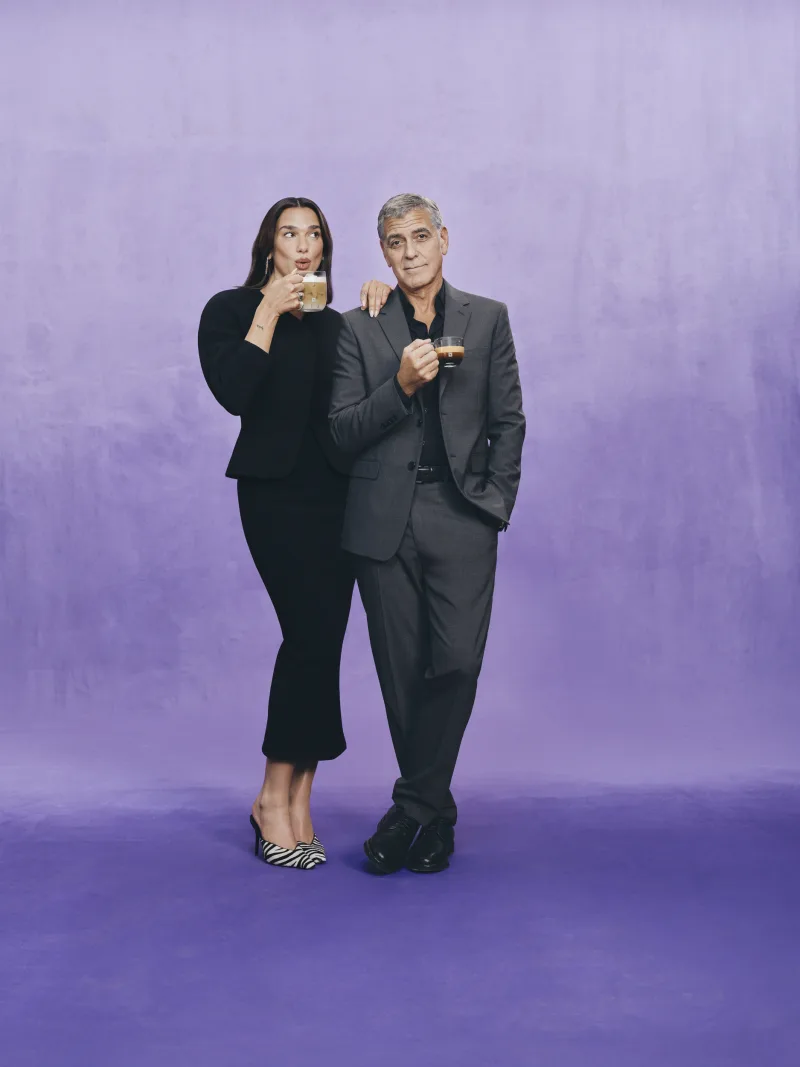 Η Dua Lipa και ο George Clooney ποζάρουν με κούπες καφέ Nespresso σε μωβ φόντο.