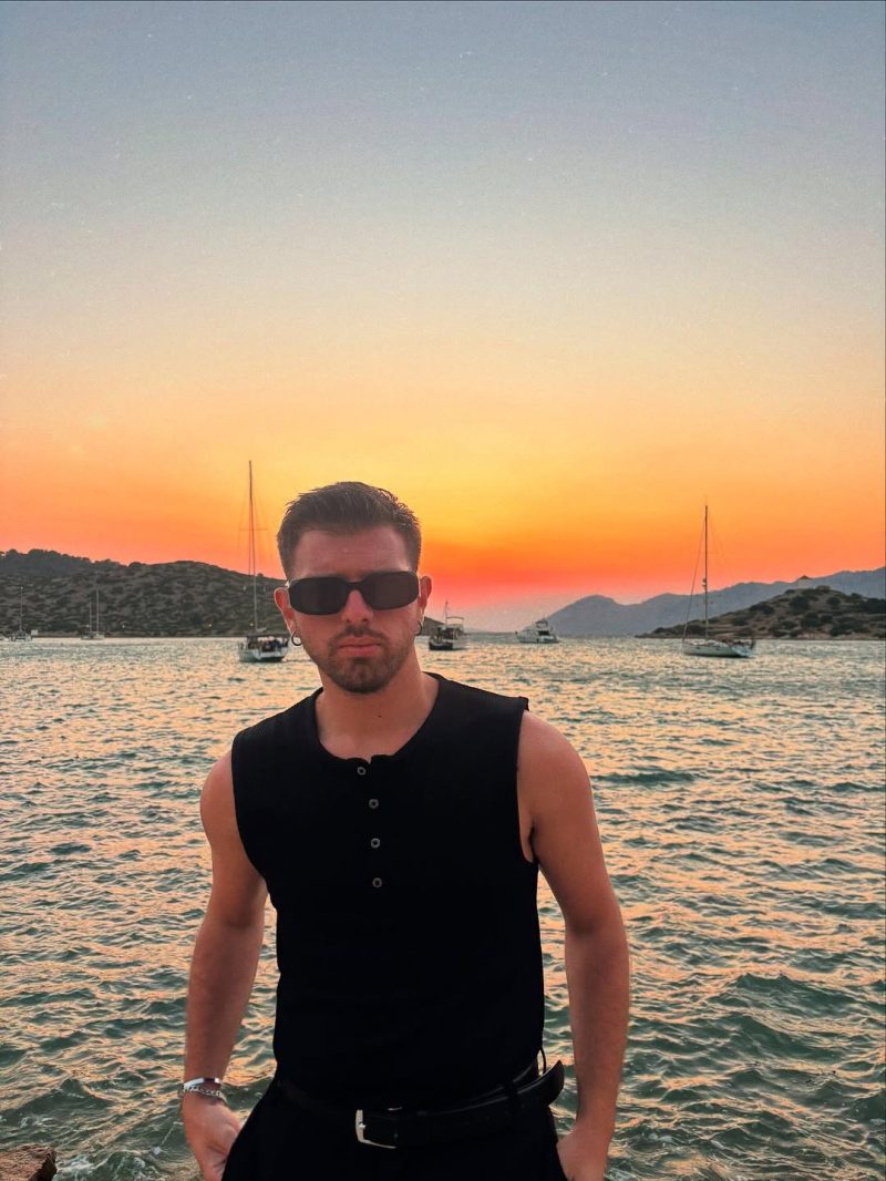 Ο creator του MAD Viral ποζάρει με γυαλιά ηλίου μπροστά σε ένα εντυπωσιακό ηλιοβασίλεμα δίπλα στη θάλασσα.