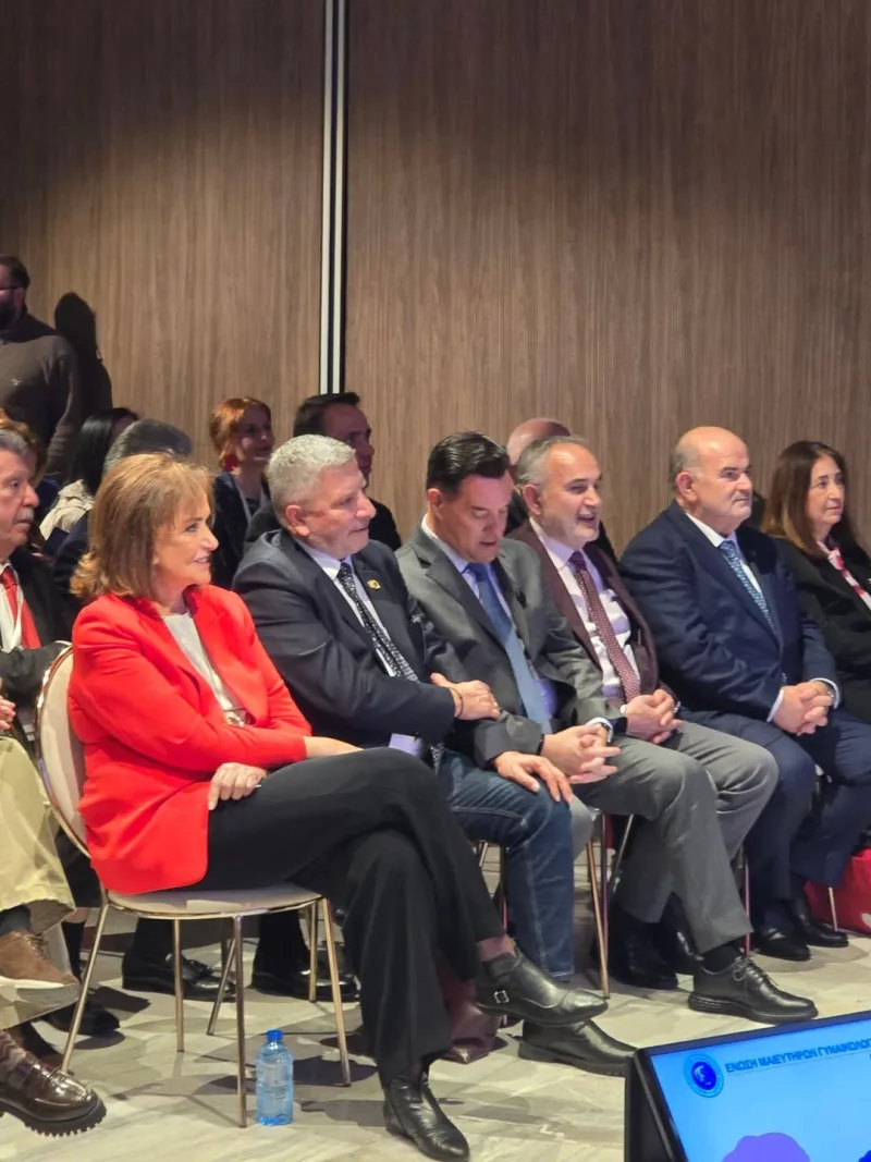 Η Ντόρα Μπακογιάννη κάθεται σε εκδήλωση, ενώ δίπλα της διακρίνονται άνδρες με κοστούμια.