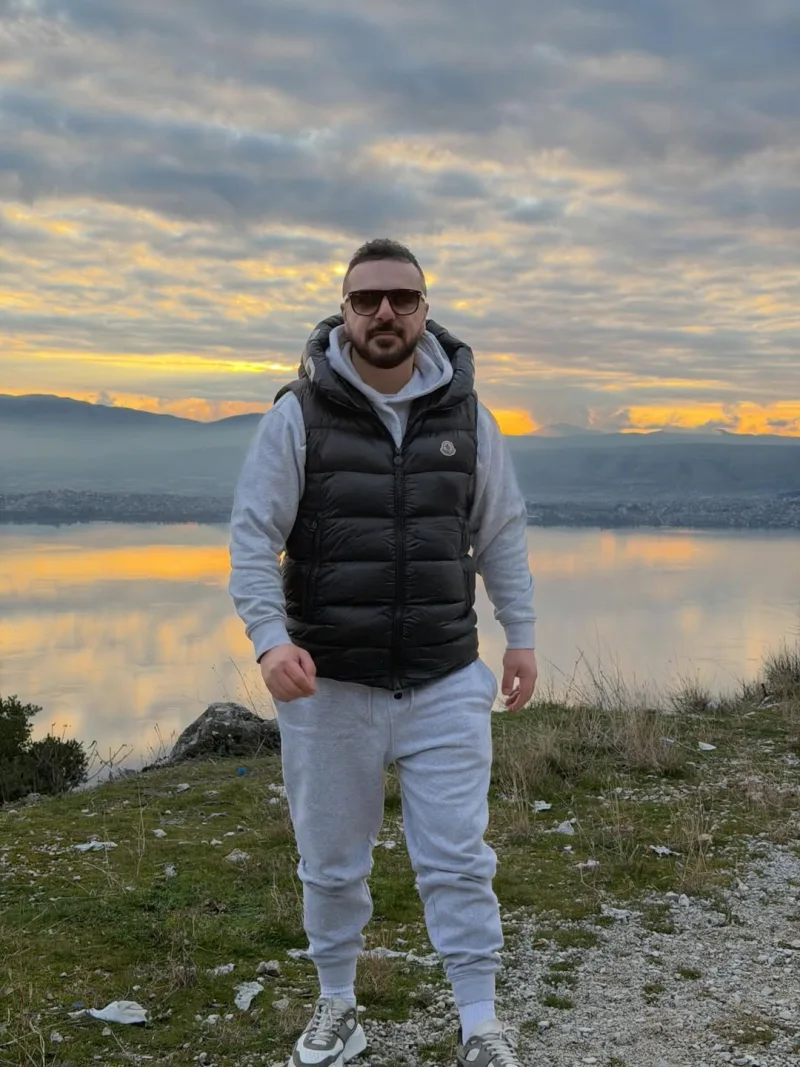 Ο Τριαντάφυλλος ποζάρει με γκρι φόρμες και γιλέκο μπροστά από τη θάλασσα με το ηλιοβασίλεμα.